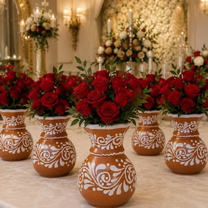 May include: A collection of terracotta vases featuring white hand-painted swirling floral patterns. Each vase has a rounded base and a flared rim, and is filled with a dense arrangement of deep red roses and green leaves, creating a uniform display.