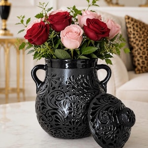 May include: A black ceramic jar with two handles and a matching lid, featuring an intricate embossed floral and scroll pattern. The jar is filled with a bouquet of deep red and soft pink roses with green leaves, creating a striking contrast.