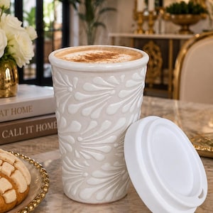 May include: A cream-colored ceramic travel mug featuring a raised, embossed floral petal pattern across its exterior. The mug is filled with a coffee beverage topped with foam and cinnamon, and it is accompanied by a separate white silicone lid.