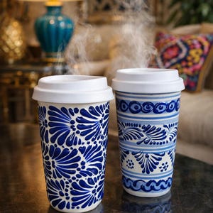 May include: Two ceramic travel mugs with white lids, emitting steam. One mug features a blue floral pattern, the other has a blue and white geometric design. The mugs are on a dark surface, suggesting a coffee or tea theme.