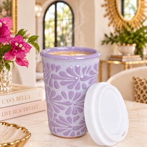 May include: A lavender ceramic travel mug with a raised floral pattern and a white silicone lid. The mug is filled with a steaming hot beverage. The exterior features a glossy finish with embossed petal-like shapes covering the surface.