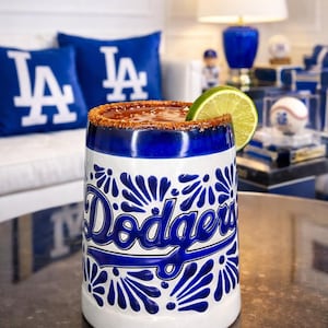 Puede incluir: Una taza de cerámica blanca y azul con el logotipo de los Dodgers de Los Ángeles y una rodaja de lima. El borde está recubierto con una especia marrón rojiza. La taza está sobre una mesa, con decoración temática de los Dodgers en el fondo.