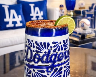 May include: A white and blue ceramic mug with the Los Angeles Dodgers logo and a lime wedge. The rim is coated with a reddish-brown spice. The mug is on a table, with Dodgers-themed decor in the background.