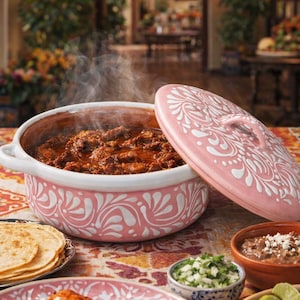 Op de afbeelding: Een roze keramische ovenschaal met een wit bloemmotief, gevuld met een stomende stoofpot. Het deksel ligt aan de zijkant. De tafel is gedekt met tortilla's, enchiladas en kleine kommen met eten, wat wijst op een Mexicaanse maaltijd.