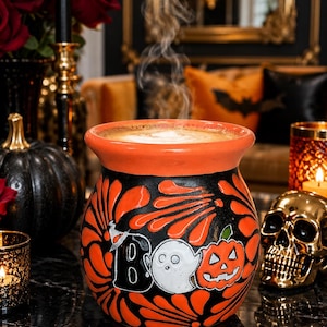May include: A round ceramic mug with a wide orange rim and a black body featuring orange floral patterns. The front displays the text "BOO" with a white ghost and an orange jack-o'-lantern pumpkin graphic. The mug is filled with a steaming beverage.