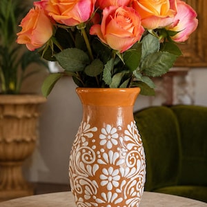 May include: A terracotta-colored ceramic vase with a flared rim and a white painted floral pattern featuring swirls and petals. The vase is filled with a bouquet of vibrant orange and pink roses with green leaves and stems extending upwards.