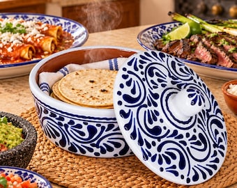 May include: A round ceramic tortilla warmer with a white base and intricate blue floral patterns. The lid is removed and placed to the side, revealing a stack of warm corn tortillas wrapped in a white cloth with blue stripes inside the container.