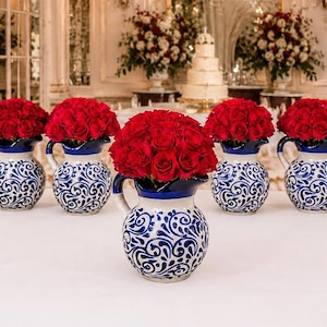 Puede incluir: Una serie de jarras de cerámica con base blanca, intrincados patrones de remolinos azules y un borde azul sólido. Cada jarra está llena con un arreglo denso y redondeado de rosas rojas vibrantes, creando una exhibición floral uniforme.