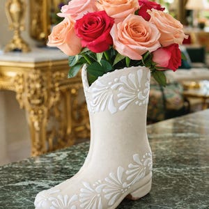 Peut inclure: Un vase en forme de botte en céramique crème, orné de motifs floraux blancs, contient un bouquet de roses rouges, roses et pêche. Le vase est posé sur une surface en marbre vert, créant une composition florale élégante.