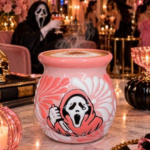 May include: A round, light pink ceramic mug featuring a raised, embossed graphic of a masked figure in a hooded robe holding a knife. The mug has a wide rim, a glossy finish, and white floral-like patterns surrounding the central character design.