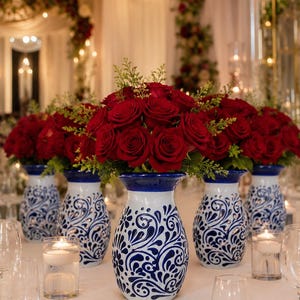 May include: Five white ceramic vases with blue hand-painted floral patterns, each filled with a dense arrangement of deep red roses and green foliage, positioned in a row on a white tablecloth with small glass candle holders in between.