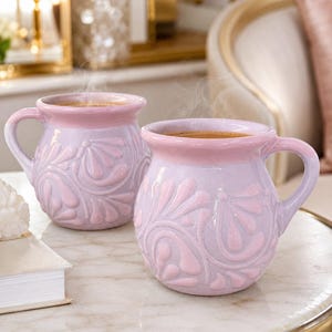 May include: Two rounded ceramic mugs with a glossy finish, featuring a light lavender body and a soft pink rim. The exterior of each mug is decorated with a raised, embossed floral pattern in a matching pink hue, and both contain a dark liquid.