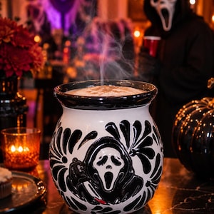 Peut inclure: Un mug en céramique blanche avec un bord noir et un motif en relief noir représentant une silhouette masquée tenant un couteau. Le mug est rempli d'une boisson chaude fumante et la surface extérieure est ornée de motifs floraux noirs.