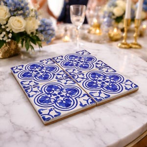 May include: Four square ceramic tiles with a blue and white floral pattern are arranged on a marble table. The tiles are a decorative element, possibly for a kitchen or dining setting. The background shows a wedding reception.