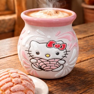May include: A ceramic mug with a pink rim and a Hello Kitty design, filled with a cappuccino. A concha pastry sits on a white plate beside the mug, all on a wooden table.