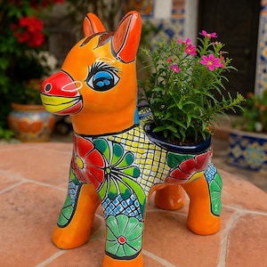 以下が含まれることがあります： 手描きのセラミックロバプランター。オレンジ色のロバは、赤、緑、青のカラフルな花柄が特徴です。プランターにはピンクの花の小さな植物が入っており、装飾的な作品に自然なタッチを加えています。