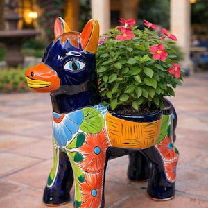 以下が含まれることがあります： 鮮やかな色で手描きされたセラミックのロバのプランター。ロバは青い体、オレンジ色のアクセント、花柄のデザインです。プランターには赤い花が付いた緑の植物が入っており、屋外の装飾に彩りを添えています。