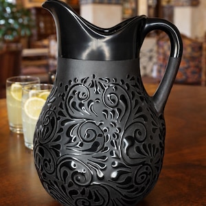 Könnte beinhalten: Eine schwarze Keramikkaraffe mit einem dekorativen Blumenmuster. Die Karaffe hat einen gebogenen Griff und einen Ausguss zum Ausgießen. Das Design ist auf den Körper der Karaffe geprägt. Die Karaffe steht auf einem Holztisch.