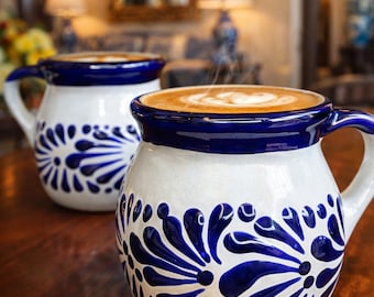 May include: Two white ceramic mugs with blue floral designs and blue rims, filled with coffee. The mugs are on a wooden table, with steam rising from the coffee. The background is blurred, suggesting a cafe setting.