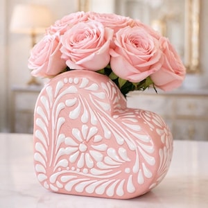 May include: A heart-shaped pink vase with white floral patterns holds a bouquet of light pink roses. The vase is on a white surface, with a blurred background featuring a mirror and a lamp.