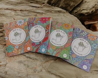 Puede incluir: Cuatro cuadernos con estampados florales en naranja, rosa, azul y verde. Cada uno presenta el texto "REM & CO" y un logotipo de regadera. Los cuadernos están sobre una superficie de piedra.
