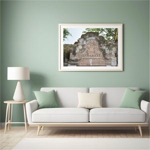 Pode incluir: Uma fotografia emoldurada de uma antiga estrutura de pedra com entalhes intrincados, contra um fundo de folhagem verde e um céu brilhante. A fotografia é exibida acima de um sofá branco com almofadas verdes e creme, criando uma cena interior serena.