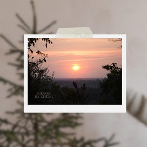 Könnte beinhalten: Fotografie eines Sonnenuntergangs über einer Landschaft, umrahmt von dunklen Silhouetten von Bäumen und Blättern. Der Himmel geht von Orange zu Rosa über, wobei die Sonne sichtbar ist. Der Text auf dem Foto lautet "PHNOM BA KHENG" und "HELLO CHRISTMAS".