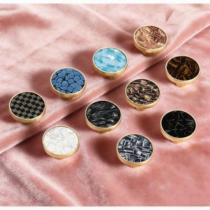 May include: A collection of decorative cabinet knobs with various mosaic and patterned designs. Each knob features a round, colorful top with gold-colored hardware. The knobs are arranged on a pink velvet surface, showcasing their diverse textures and colors.