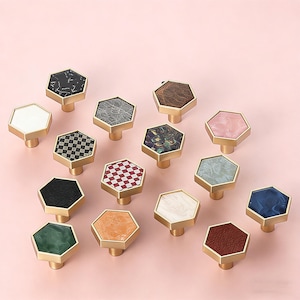 May include: A collection of decorative cabinet knobs with hexagonal faces. The knobs feature various patterns and colors, including marble, checkered, and solid hues, with gold-colored bases. The knobs are arranged on a pink background.