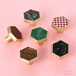 May include: A collection of six hexagonal cabinet knobs with brass bases. The knobs feature various decorative tops, including a red and white checkered pattern, a green marble effect, and a brown wood grain.