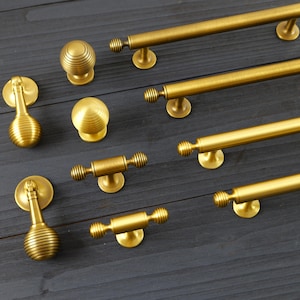 May include: A collection of gold-toned cabinet hardware, including knobs and pulls. The knobs have a round, ribbed design, while the pulls are bar-shaped with decorative ends. The hardware is arranged on a dark wooden surface, showcasing various sizes and styles.