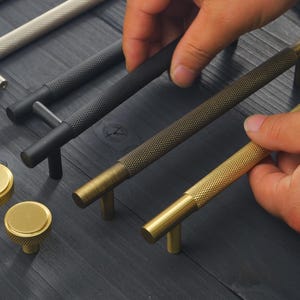 May include: Close-up of various cabinet hardware, including gold and black textured bar pulls and gold knobs. The hardware is displayed on a dark wood surface, showcasing the different finishes and designs. The bar pulls are being held by a person.