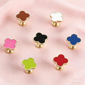 Può includere: Una collezione di pomelli decorativi per mobili con un design a trifoglio. Ogni pomello presenta una base color oro e una parte superiore a trifoglio a quattro foglie in vari colori, tra cui marrone, bianco, rosa, nero, blu, verde e rosso.