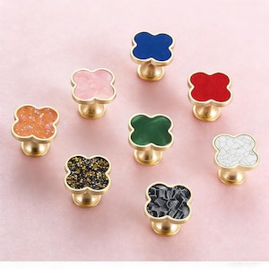May include: A collection of eight decorative cabinet knobs. Each knob features a four-leaf clover design with a gold-colored base. The clover tops come in various colors and textures, including pink, blue, red, green, orange, black, and white.