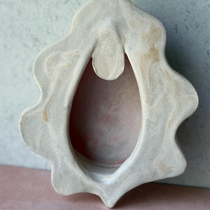May include: A cream-colored, ceramic wall hanging with an organic, scalloped shape. The central opening is oval, and the piece has a textured surface with subtle variations in color. The design is reminiscent of a stylized flower or abstract form.