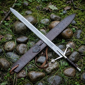 May include: A double-edged steel sword with a central fuller and a cross-shaped guard featuring four-lobed terminals. The handle is wrapped in dark material, and the sword is accompanied by a dark brown leather scabbard with a leather strap.