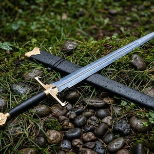 May include: A longsword with a double-edged steel blade featuring a central fuller, a black leather-wrapped grip, and a gold-coloured metal crossguard and pommel. The sword is accompanied by a black scabbard with gold-coloured metal fittings.