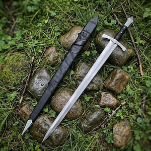 Puede incluir: Una espada de acero de doble filo con una guarda plateada y un mango negro, colocada junto a una vaina de cuero negro a juego con una punta de metal y un detalle de cordón de cuero envuelto, sobre piedras cubiertas de musgo y hierba.