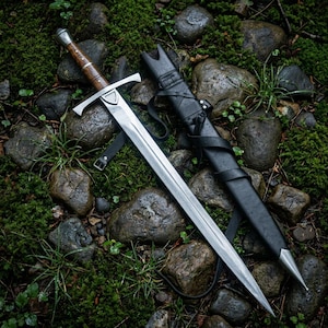 May include: A steel sword with a wooden handle and a silver pommel, featuring a crossguard with a central shield detail. The sword is accompanied by a black leather scabbard with silver metal fittings, including a pointed tip and straps.