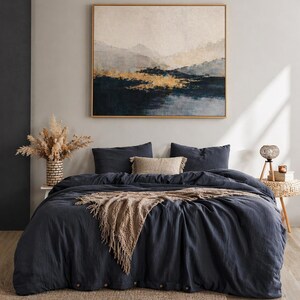 Könnte beinhalten: Schlafzimmer-Interieur mit einer dunkelblauen Bettdecke und Kissen, einer beigefarbenen Decke mit Fransen und einem gewebten Teppich. Ein gerahmtes Landschaftsbild hängt über dem Bett. Eine Korbbas mit getrockneten Pflanzen und ein kleiner Beistelltisch vervollständigen die Einrichtung.
