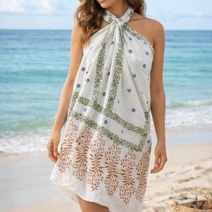 Könnte beinhalten: Weißes Neckholder-Kleid mit floralem und geometrischem Muster in Braun-, Grün- und Blautönen. Das Kleid wird an einem Strand mit dem Meer im Hintergrund getragen. Das Kleid ist etwa 140 cm lang.