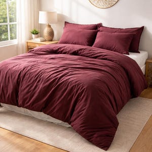 Könnte beinhalten: Ein Bett mit einer weinroten Bettdecke und passenden Kissenbezügen. Das Bett ist gemacht und bezugsfertig. Auf dem Boden liegt ein beiger Teppich. Eine dekorative Wanddekoration und eine Lampe befinden sich ebenfalls im Raum.