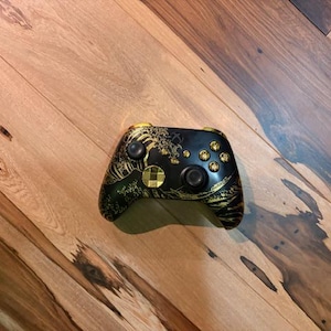 Puede incluir: Mando de Xbox negro con un diseño de olas doradas y verdes. El mando tiene detalles dorados en los botones y joysticks. El diseño está inspirado en el arte japonés.