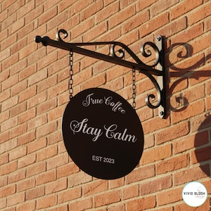 May include: A black, oval-shaped sign for a coffee shop, hanging from a decorative metal bracket. The sign reads "True Coffee Stay Calm EST 2023" in white script. The sign is suspended by chains against a brick wall.