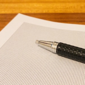 May include: A black pen with a textured grip rests on a sheet of graph paper. The pen has a silver tip and accents. The paper is white with a grid pattern, and the background is a wooden surface.