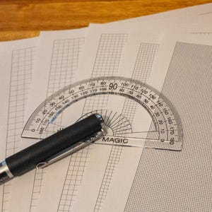 May include: A clear plastic protractor with degree markings rests on graph paper, alongside a black pen. The word "MAGIC" is printed on the protractor. The pen has a silver clip and the word "HEMA" is printed on the pen.