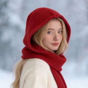 May include: A red knit hood and scarf set. The hood is pulled up, framing the face, and the scarf is wrapped around the neck. The set is made of a thick, textured knit material. The person is wearing a cream-colored sweater.