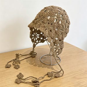 Può includere: Una cuffia all'uncinetto color marrone chiaro caratterizzata da un motivo floreale ripetuto su tutto il tessuto. Il copricapo include lunghi lacci pendenti su ciascun lato, rifiniti con piccoli dettagli a fiore a quattro petali alle estremità.