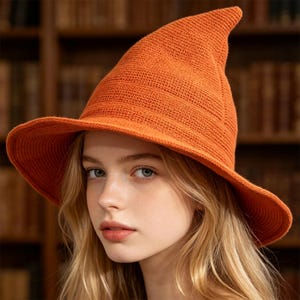May include: A bright orange, pointed witch hat crafted from a textured, knit fabric. The hat features a wide, slightly drooping brim and a tall, conical crown that tapers to a rounded tip, showcasing a consistent, dense stitch pattern throughout.