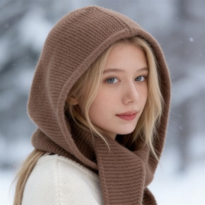 May include: A brown knitted hood and scarf set. The hood is designed to cover the head and neck, with a scarf that drapes down the front. The set is made of a soft, textured knit material. The background is a snowy, winter scene.